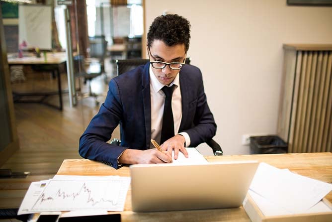 Man intelligent economist writing notary in textbook during work on portable laptop computer. Male creating new business plan sitting at table with netbook and paper documents with financial charts.