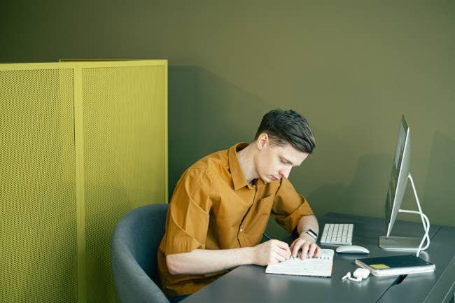 Man writing in notebook sitting in front of computer