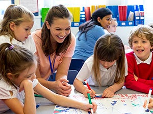 A classroom of young boys and girls drawing on one large piece of paper. The children are wearing school uniform while their teacher watches.