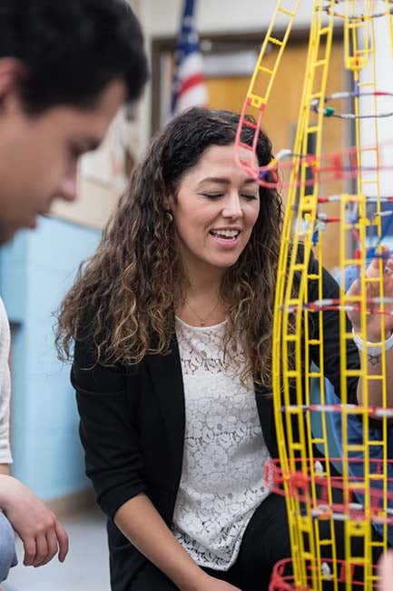 An art teacher kneels down and talks with her surrounding students about the form and shape of the colorful yellow sculpture they're studying.