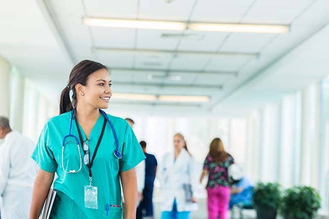 Confident young Hispanic nurse looks to the side while walking in a hospital corridor. She is wearing scrubs, stethoscope, glasses and id badge. She is carrying medical charts. Medical professionals are walking in the background.