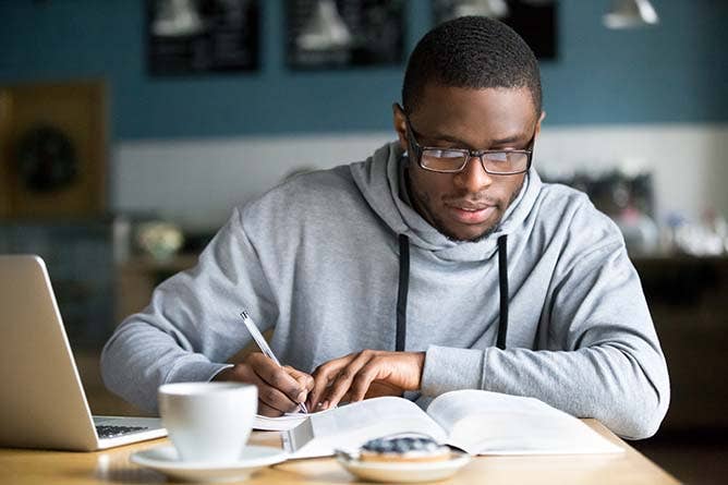 Focused student in glasses making notes writing down information from book in cafe preparing for test or exam, young serious man studying or working in coffee house
