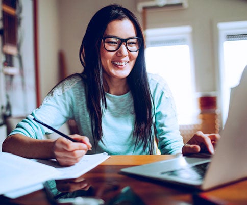 Woman wearing glasses writing on a paper next to her laptop