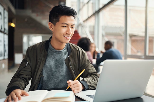 Young adult man looking at computer and smiling while writing with pencil in hand and notebook
