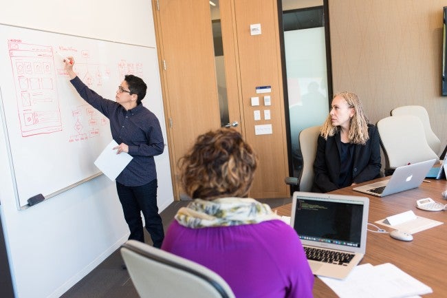 Two women watching a third person present information on a whiteboard