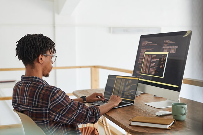 Man writing code at  laptop with large monitor behind it