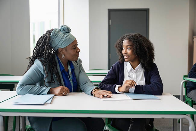 Front view of supportive female teacher in early 50s sitting at desk with teenage schoolgirl while discussing her writing assignment.