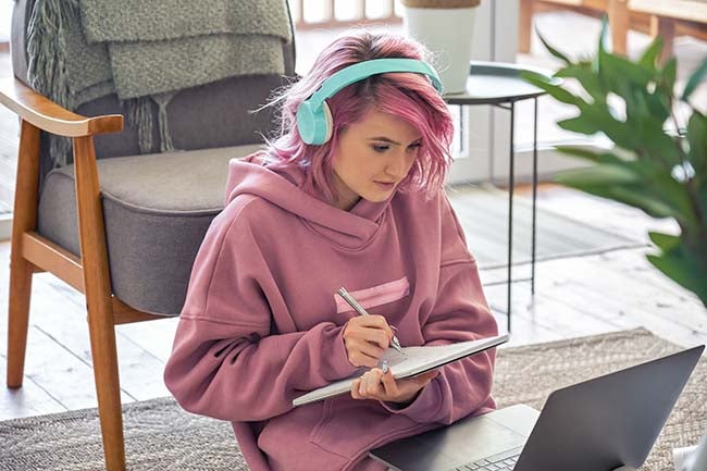 Focused teen girl school college student pink hair wear headphones write notes watching webinar online video conference calling on laptop computer sit on floor working learning online at home.