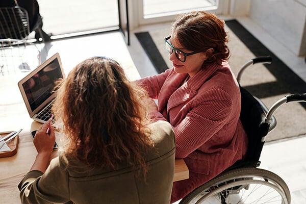 Person using a wheelchair positioned next to a co-worker. Both are working on a laptop together.